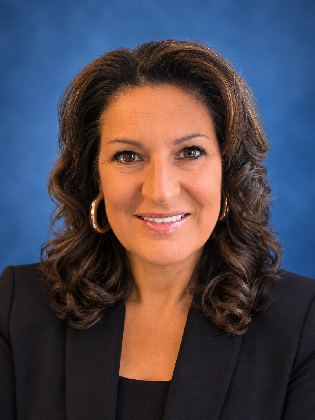 A professional headshot of a smiling woman with dark wavy hair and a dark blazer, set against a blue background.